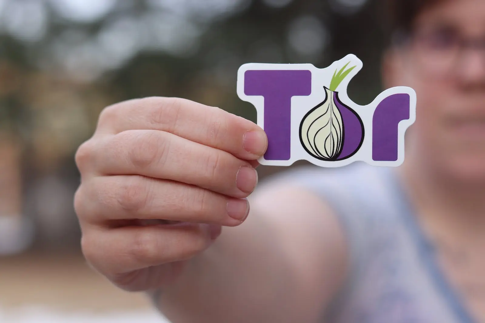 Een hand houdt een sticker vast met het Tor-projectlogo, een paarse letter 'T' en een ui-afbeelding