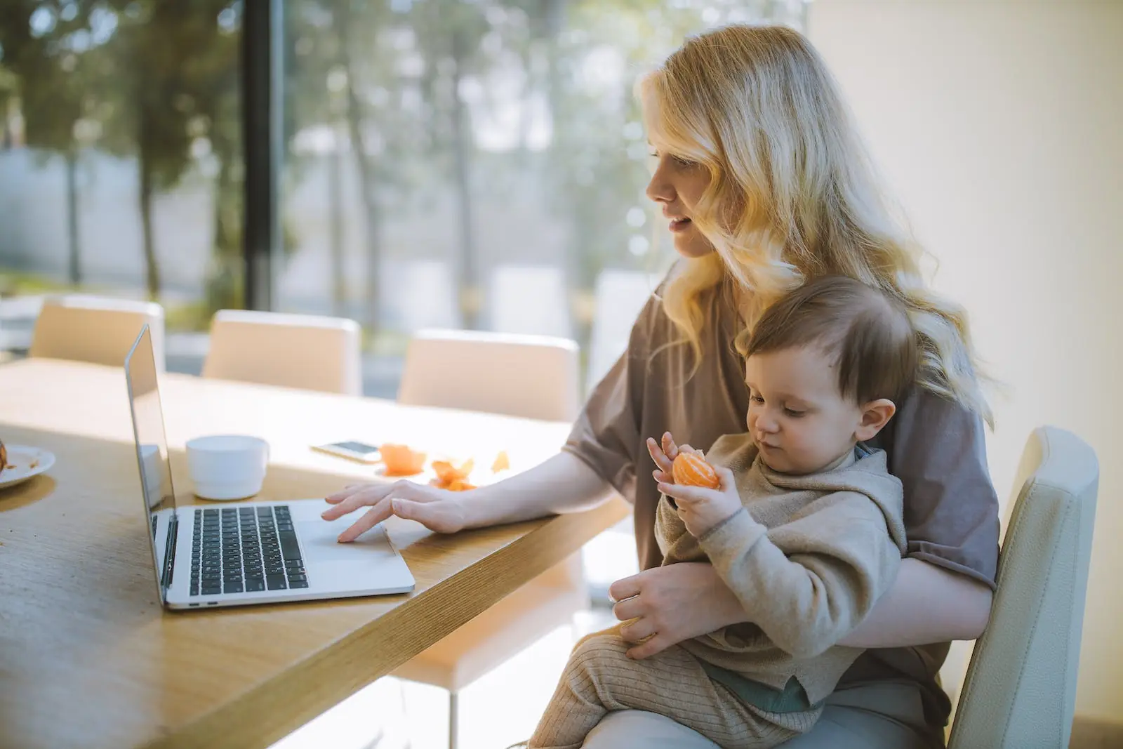 Een vrouw die aan een tafel zit en een laptop gebruikt, terwijl ze een jong kind op schoot houdt dat met een stuk fruit speelt