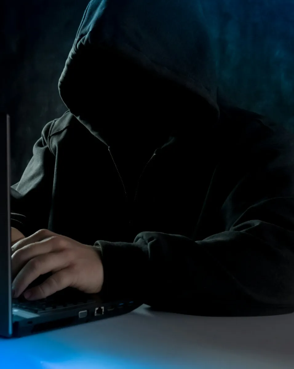 Onherkenbare persoon in donkere hoodie typt op laptop in schemerige kamer.