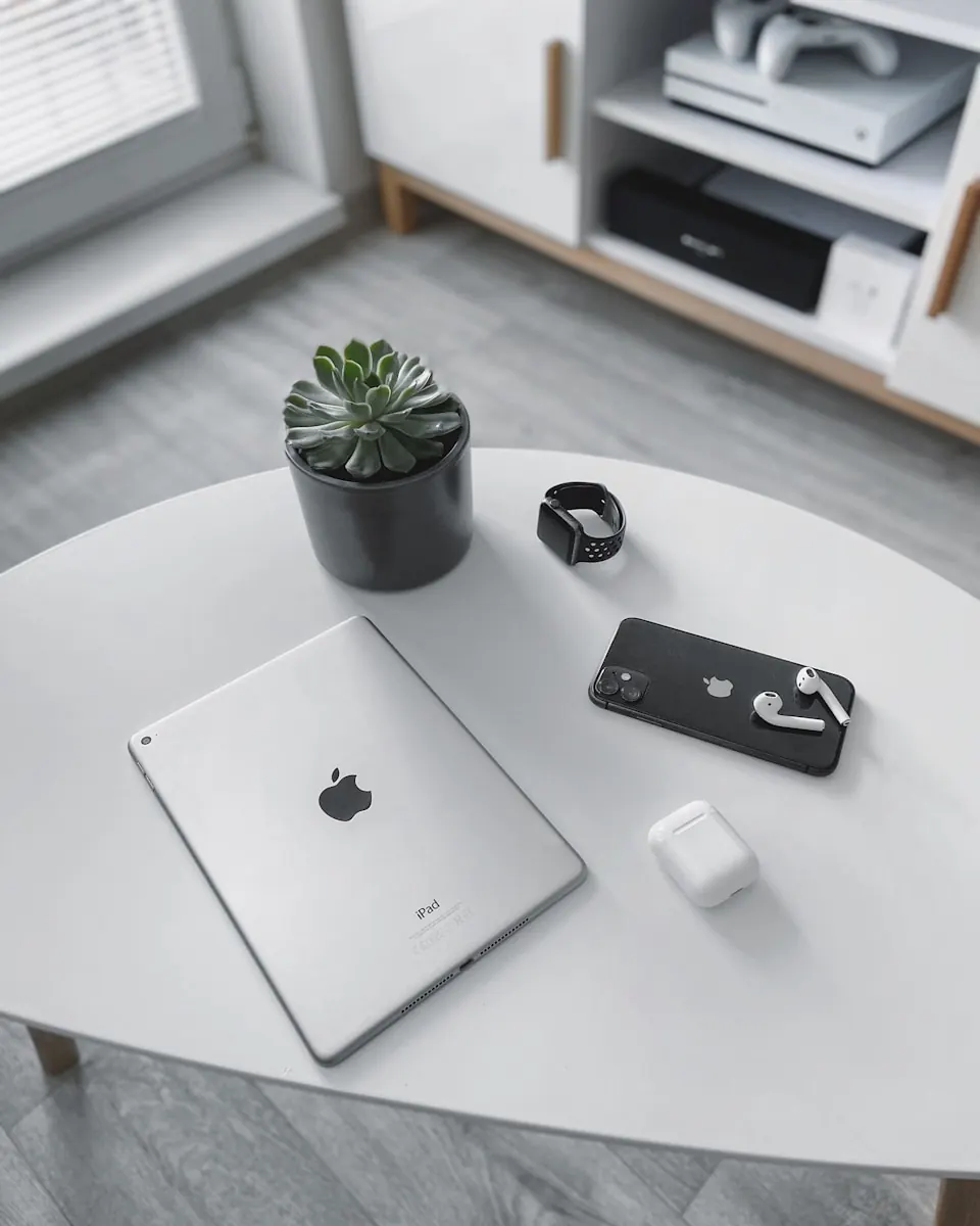 Een minimalistisch ingerichte kamer met een witte ronde tafel waarop een iPad, een zwarte smartwatch, een iPhone met AirPods, en een pot met een vetplantje staan.